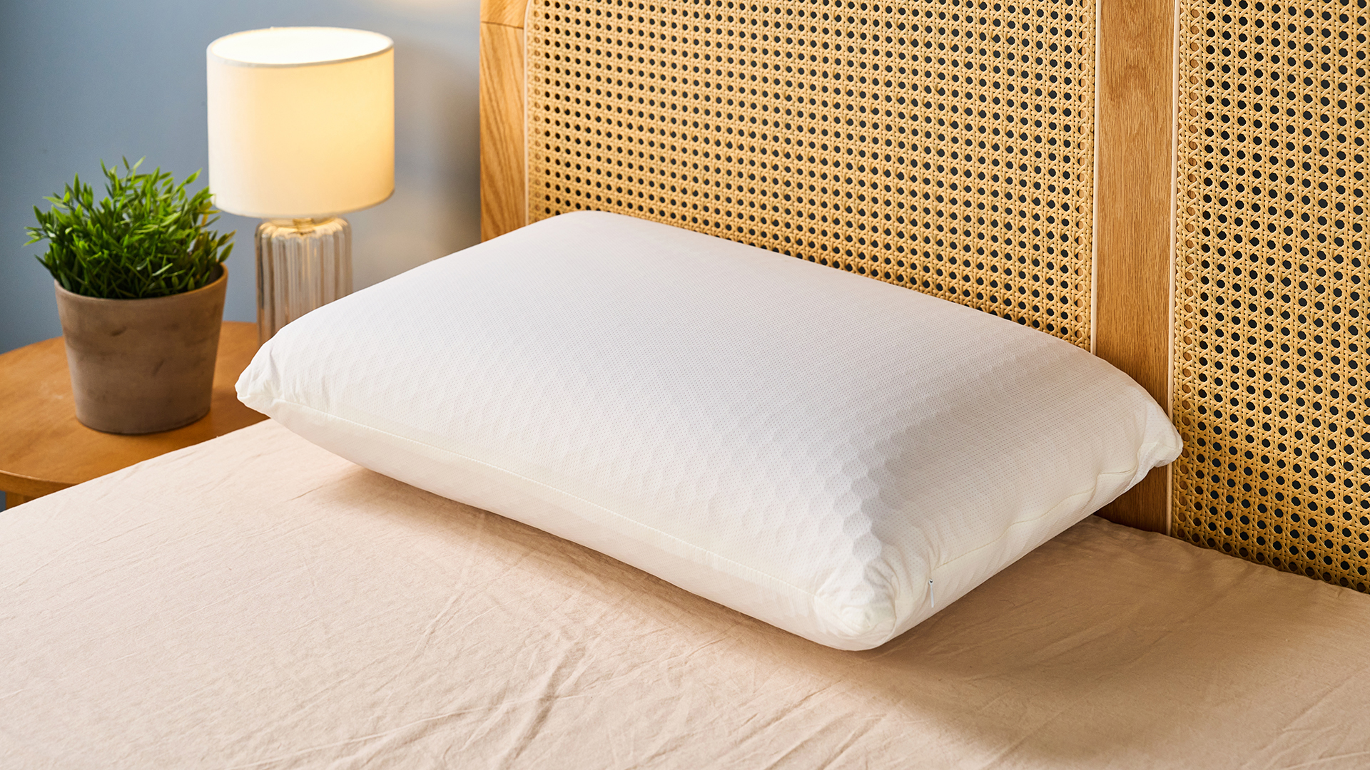 Το Purple Harmony Pillow κάθεται στο κεφάλι ενός στρώματος σε ένα σκελετό κρεβατιού, στα αριστερά ένα κομοδίνο με μια γλάστρα και ένα φωτιστικό