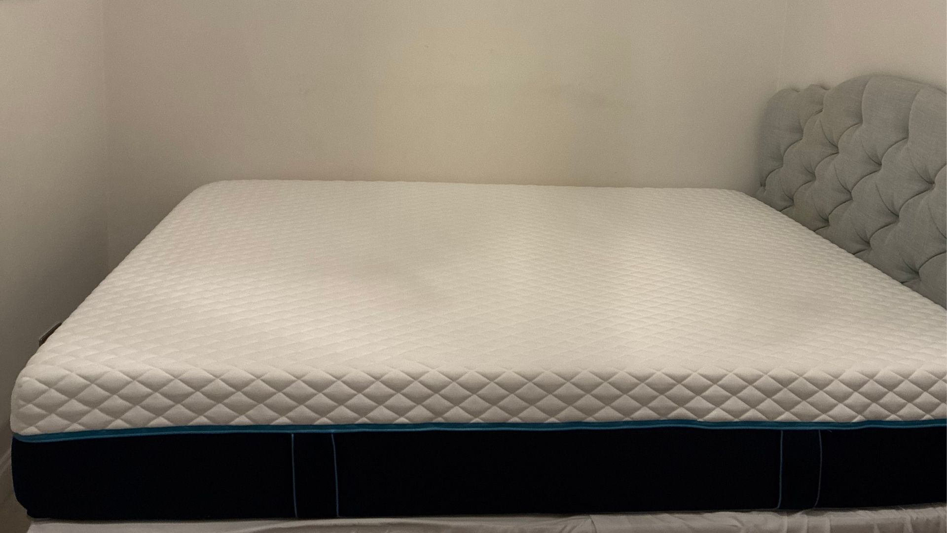 The image shows a white mattress with dark blue sides placed on a mattress base with a grey headboard, set inside a white bedroom