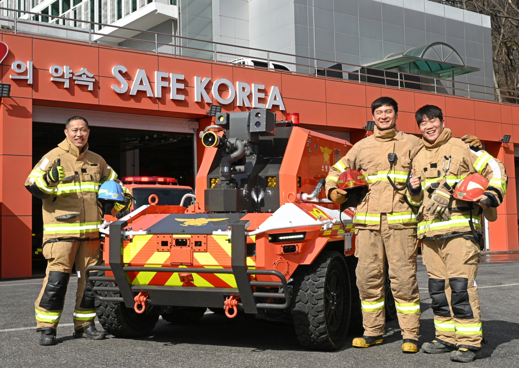 τρεις πυροσβέστες τρίβουν με ένα κόκκινο και κίτρινο πυροσβεστικό ρομπότ έξω από έναν πυροσβεστικό σταθμό