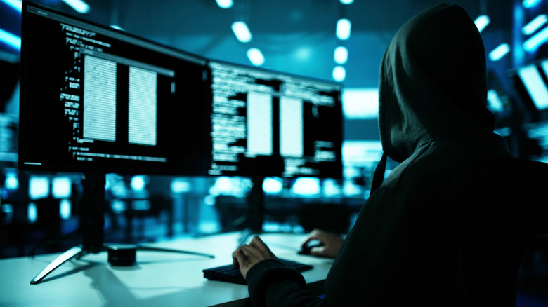 Ένας χάκερ που φοράει κουκούλα ενώ κάθεται σε ένα γραφείο και κοιτάζει τον κώδικα σε δύο οθόνες υπολογιστών